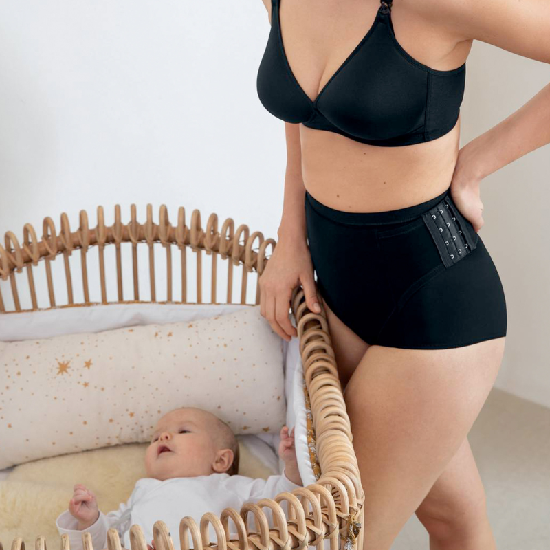 woman body in an anita black nursing bra and anita black rebelt standing next to a crib with a baby in it (representing post-partum products and care)